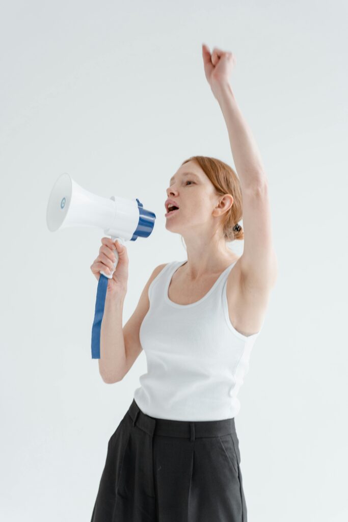 Woman raising arm and shouting into megaphone indoors, white background.