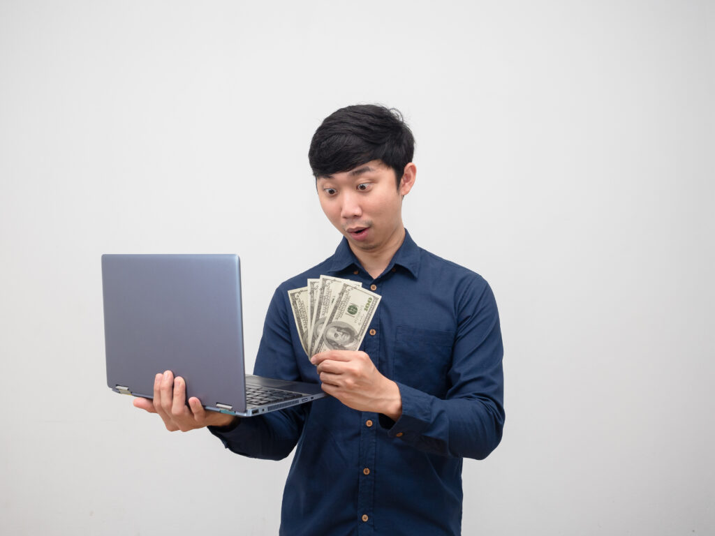 Digital creator look money in his hand and holding laptop feeling amazed on white background